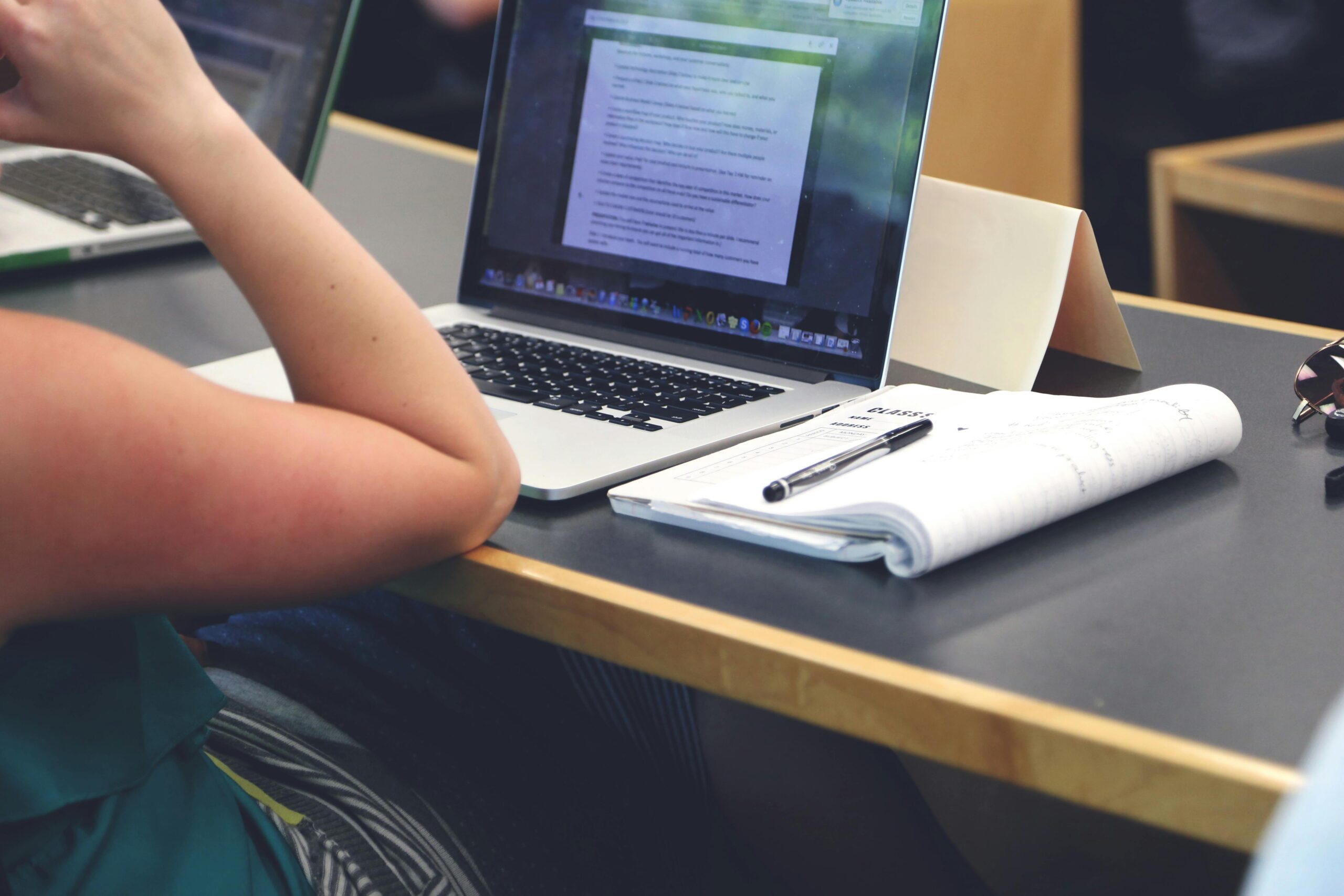 A student in a classroom setting using a laptop and taking notes in a notebook.