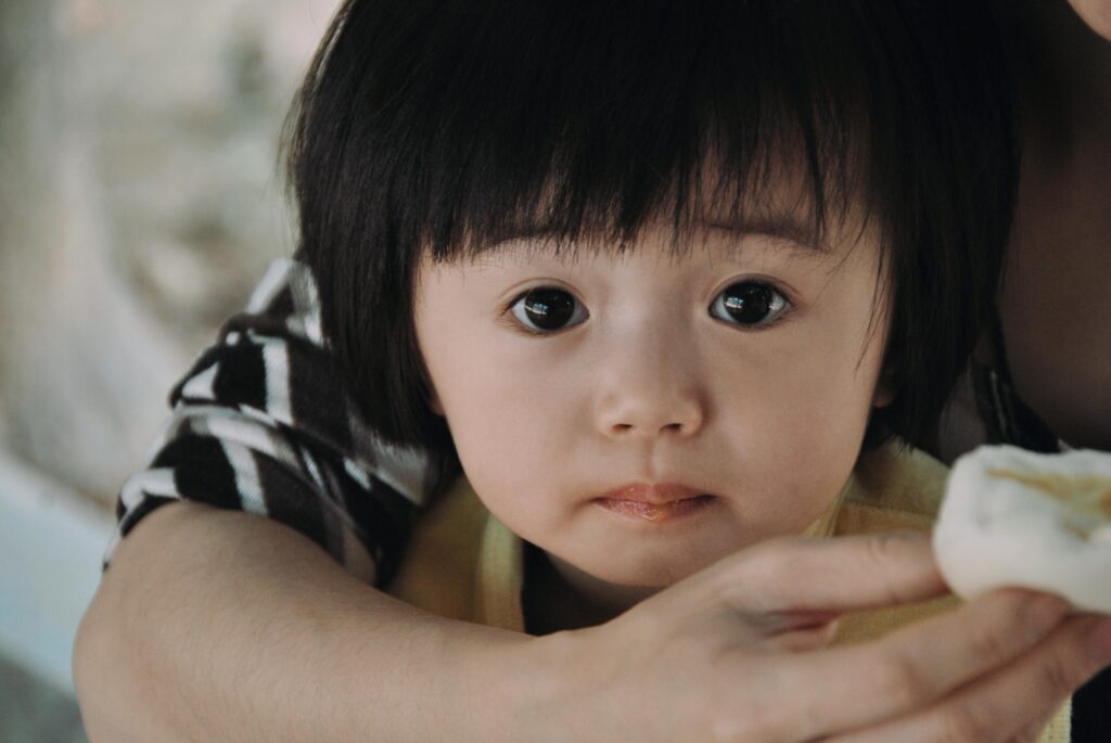 Cute Asian child in parent's arms, holding a snack, with an expressive gaze.