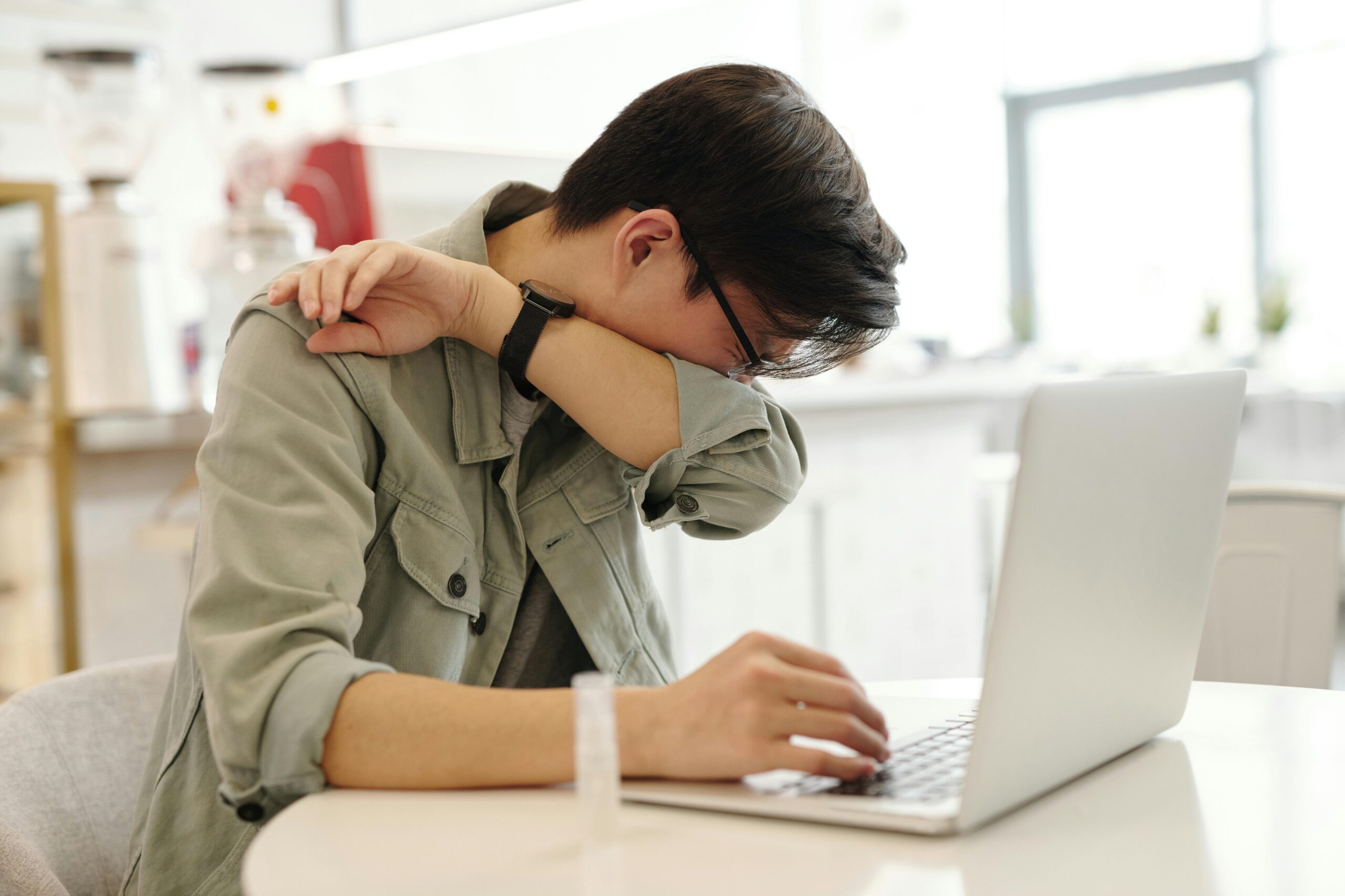 A man sneezes into his elbow while using a laptop, depicting common cold symptoms in a modern workspace.