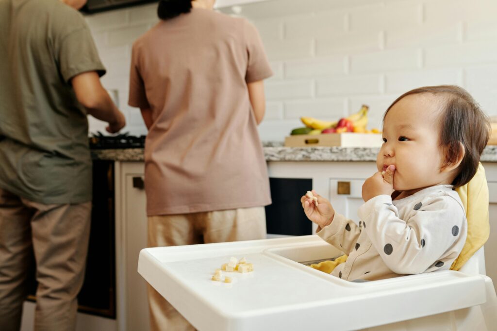 Cute toddler sits in a high chair enjoying a meal, while parents cook in the background of a modern kitchen.