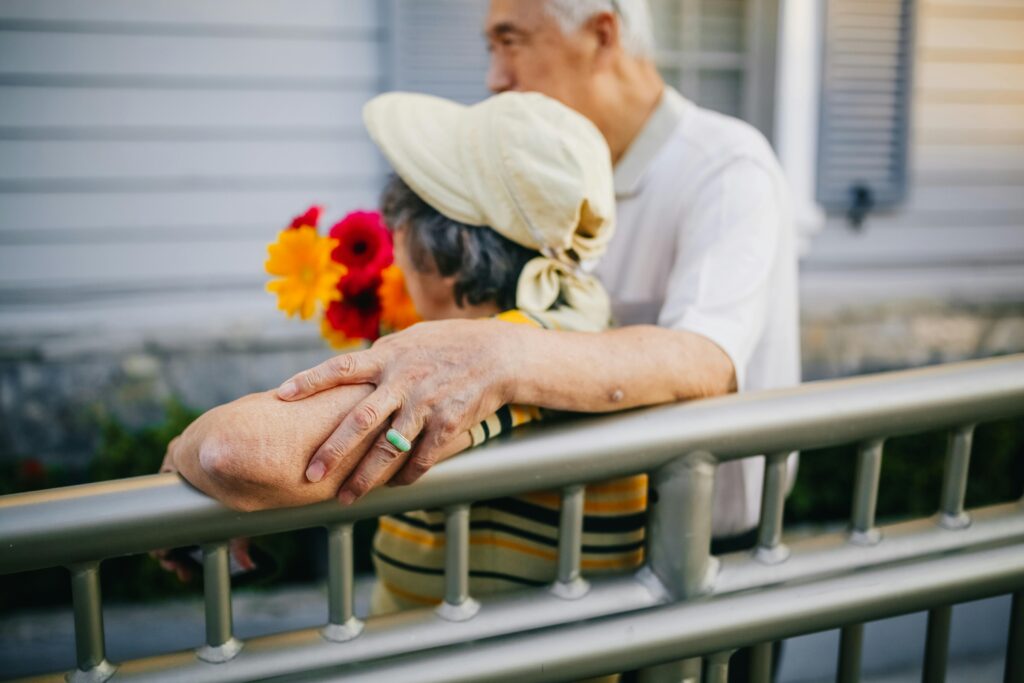 Warm moment of an elderly Asian couple embracing near a fence, holding a colorful bouquet outdoors.